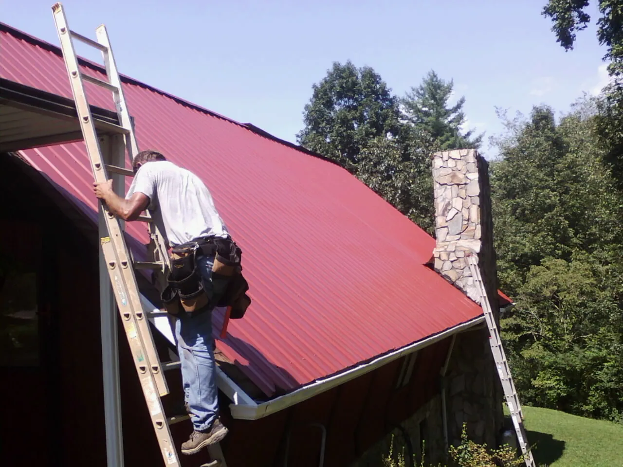 Roofer on a ladder beside a red metal roof and stone chimney — Soffit & Fascia Repair by Roofing Brothers in Gotha
